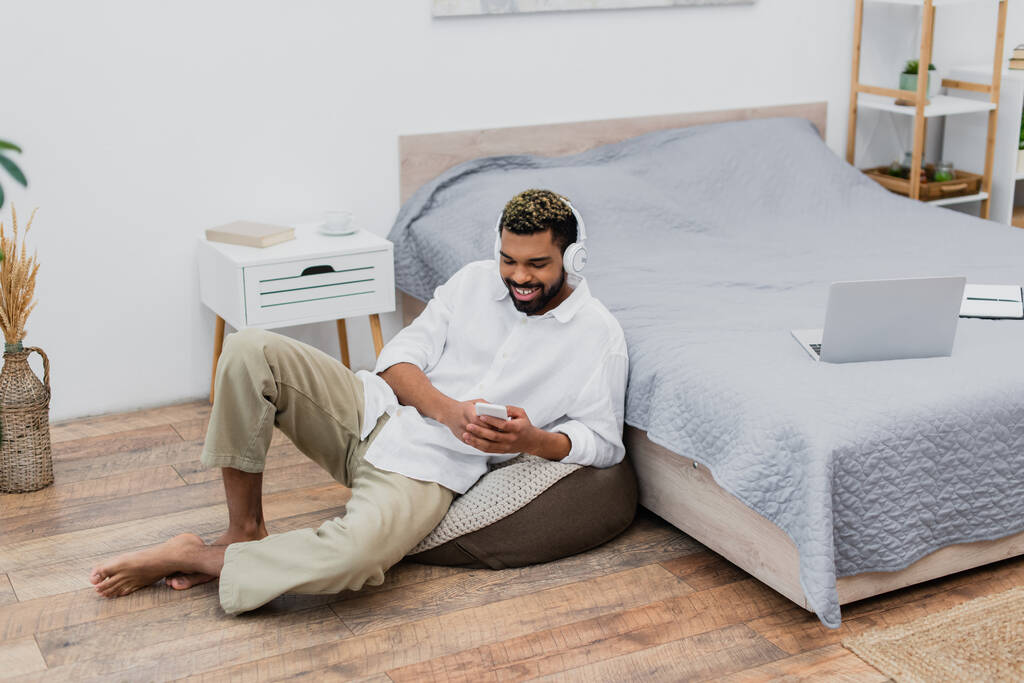 homem americano africano feliz em fones de ouvido sem fio usando smartphone enquanto sentado perto da cama - Foto, Imagem