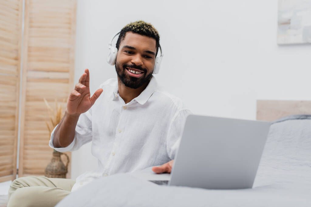 homem americano africano feliz em fone de ouvido sem fio acenando mão durante chamada de vídeo no laptop  - Foto, Imagem