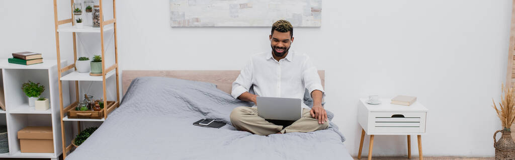 allegro uomo africano americano in auricolari senza fili utilizzando il computer portatile mentre seduto sul letto, banner - Foto, immagini