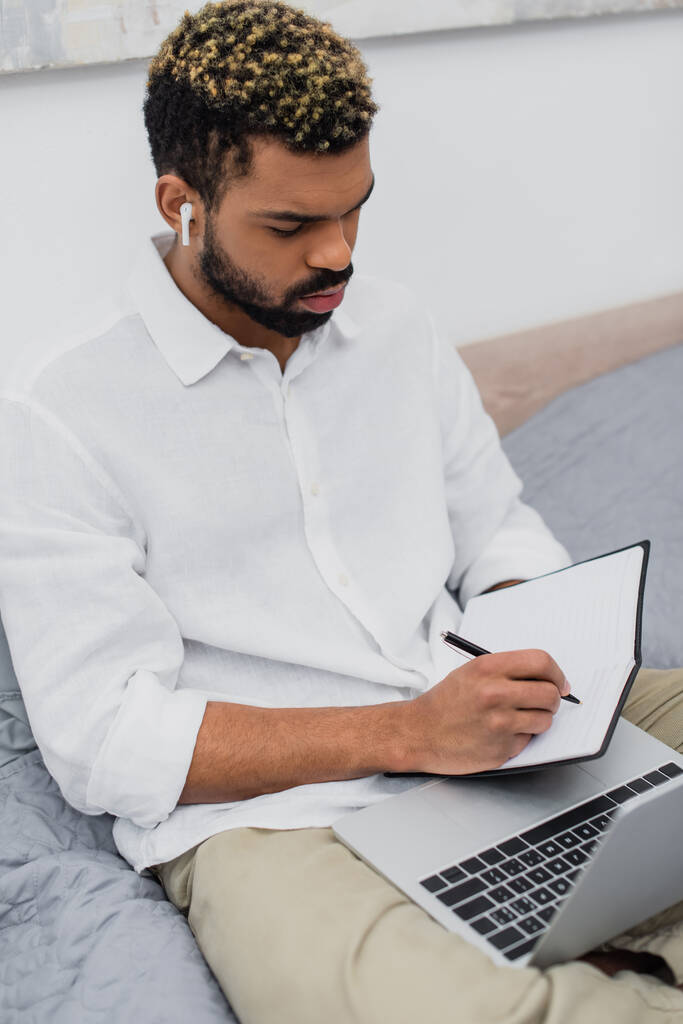 joven afroamericano hombre en auriculares inalámbricos haciendo notas cerca de la computadora portátil en el dormitorio  - Foto, Imagen