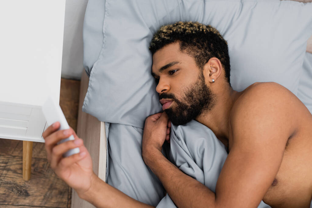 vista superior del hombre afroamericano barbudo usando un teléfono inteligente mientras está acostado en la cama por la mañana  - Foto, Imagen
