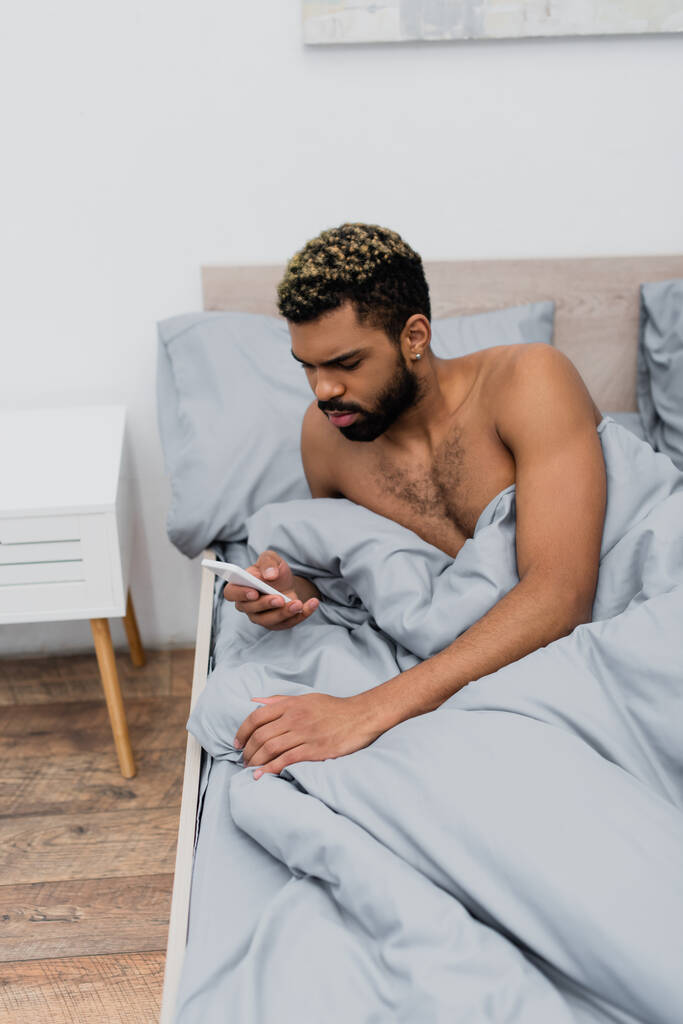 bez koszulki Afroamerykanin człowiek z farbowane włosy wiadomości na smartfon w łóżku - Zdjęcie, obraz