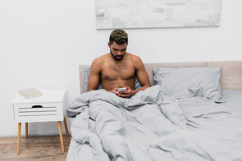 hombre afroamericano sin camisa con el pelo teñido charlando en el teléfono inteligente en el dormitorio  - Foto, imagen