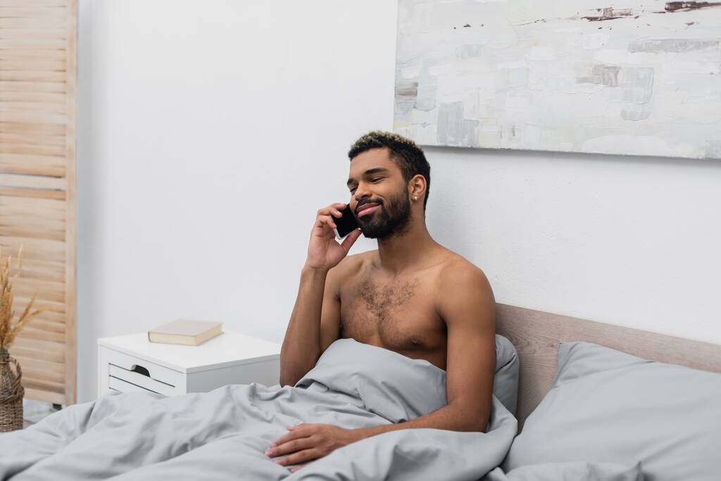 bez koszulki i szczęśliwy Afrykański Amerykanin z farbowanymi włosami rozmawiający przez telefon komórkowy w łóżku - Zdjęcie, obraz