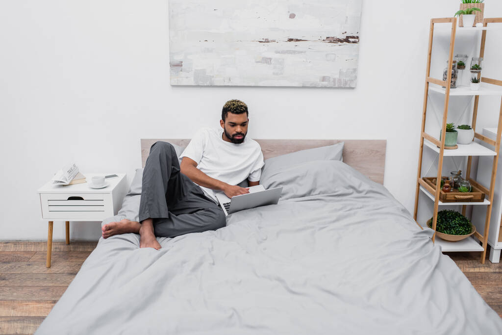 Afrikanisch-amerikanischer Mann mit gefärbten Haaren benutzt Laptop und arbeitet von zu Hause aus im Bett - Foto, Bild