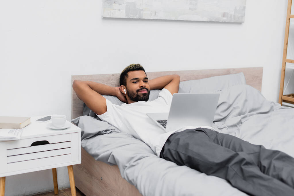 glücklich afrikanisch-amerikanischer Mann in drahtlosem Kopfhörer Film anschauen, während er mit Laptop im Bett liegt  - Foto, Bild