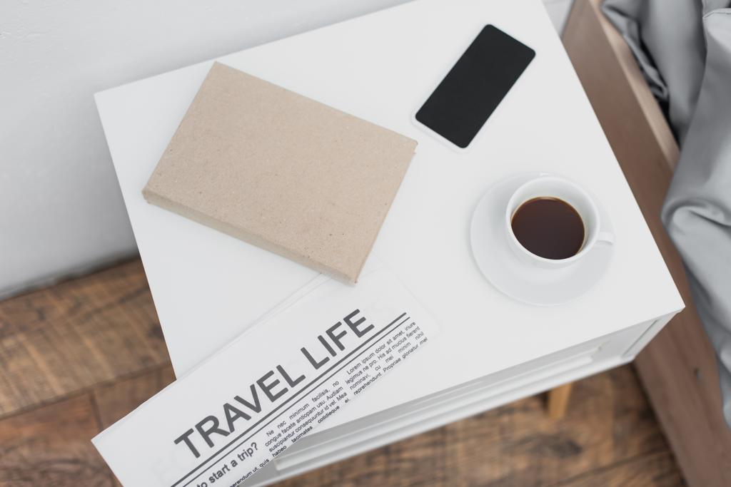 top view of cup with coffee and smartphone near newspaper with travel life lettering and book on white bedside table - Photo, Image