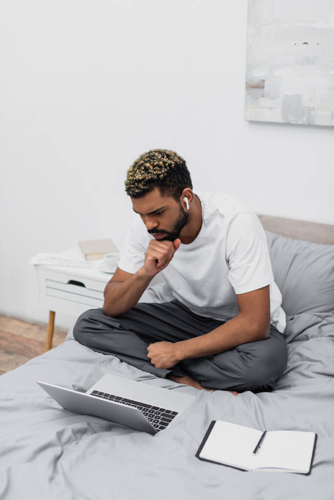 nachdenklicher afrikanisch-amerikanischer Mann mit drahtlosem Kopfhörer, der Laptop benutzt und von zu Hause aus im Bett arbeitet - Foto, Bild