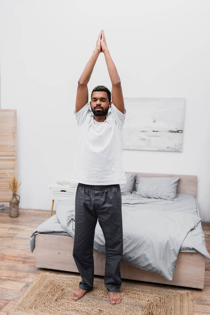 bärtiger afrikanisch-amerikanischer Mann mit geschlossenen Augen steht in Baum-Yoga-Pose im Schlafzimmer  - Foto, Bild