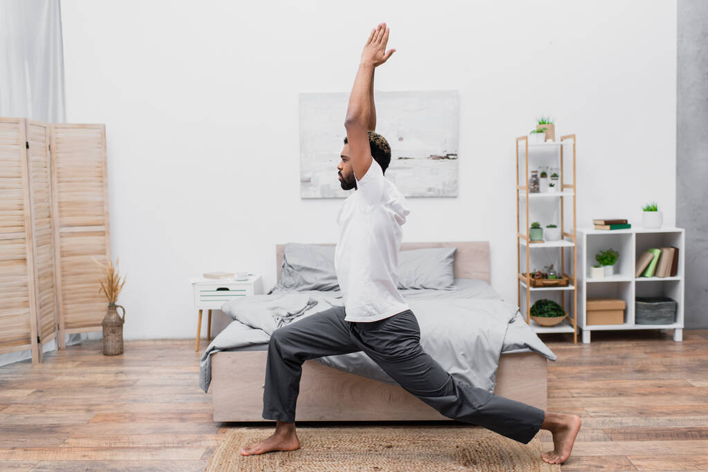 vista lateral do homem americano africano barbudo praticando ioga em pose guerreira e meditando no quarto  - Foto, Imagem