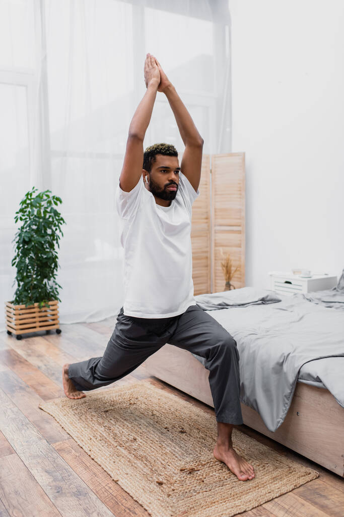 hombre afroamericano barbudo practicando yoga en pose guerrera y meditando en dormitorio moderno  - Foto, imagen