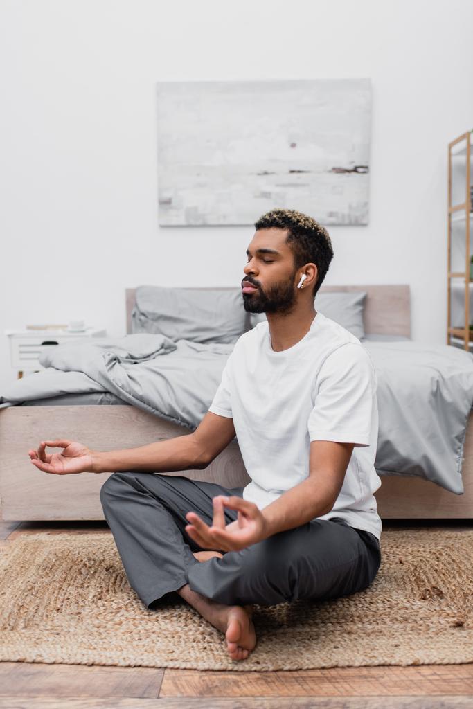 hombre afroamericano barbudo en auricular inalámbrico meditando con los ojos cerrados mientras está sentado cerca de la cama en casa - Foto, Imagen