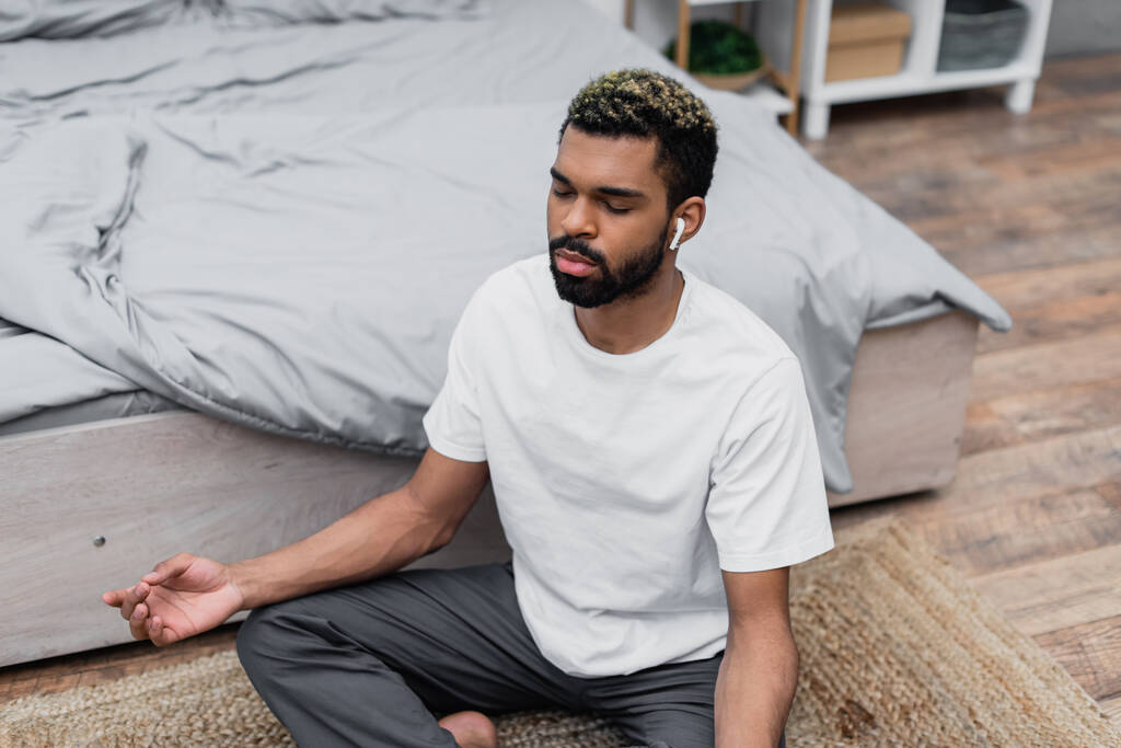 vue grand angle de barbu homme afro-américain méditant avec les yeux fermés tout en étant assis près du lit à la maison - Photo, image