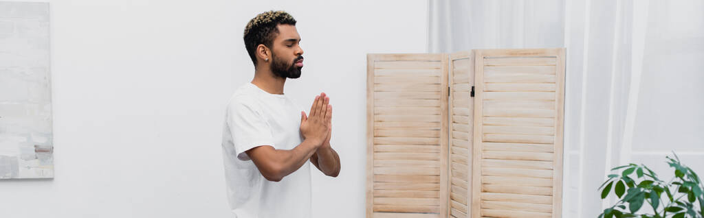 barbudo hombre afroamericano meditando con las manos de oración cerca de la pantalla plegable, pancarta - Foto, Imagen