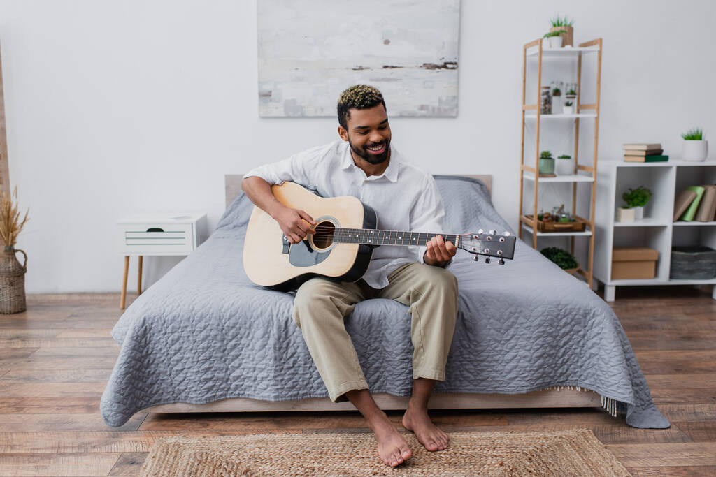 uomo afroamericano scalzo e felice con capelli tinti e barba suonare la chitarra acustica in camera da letto - Foto, immagini
