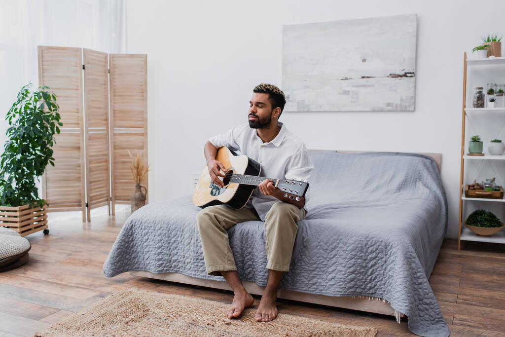 boso Afroamerykanin człowiek z farbowanymi włosami i brodą gra na gitarze akustycznej w sypialni - Zdjęcie, obraz
