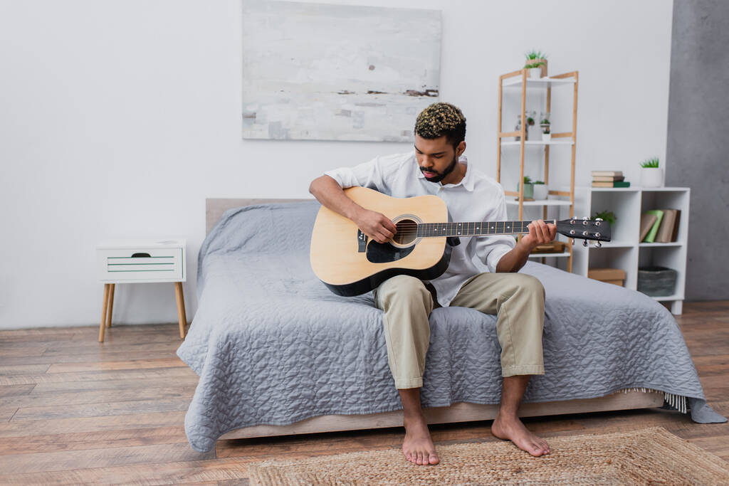 barefoot jonge Afrikaanse Amerikaanse man met geverfd haar en baard spelen akoestische gitaar in de slaapkamer - Foto, afbeelding