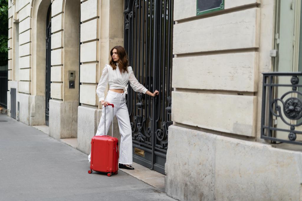 Modische Frau mit Koffer steht in der Nähe eines Gebäudes an der Stadtstraße in Paris  - Foto, Bild