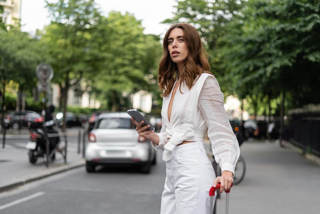 Стильная женщина с сотовым телефоном и чемоданом, стоящая возле дороги на улице в Париже  - Фото, изображение