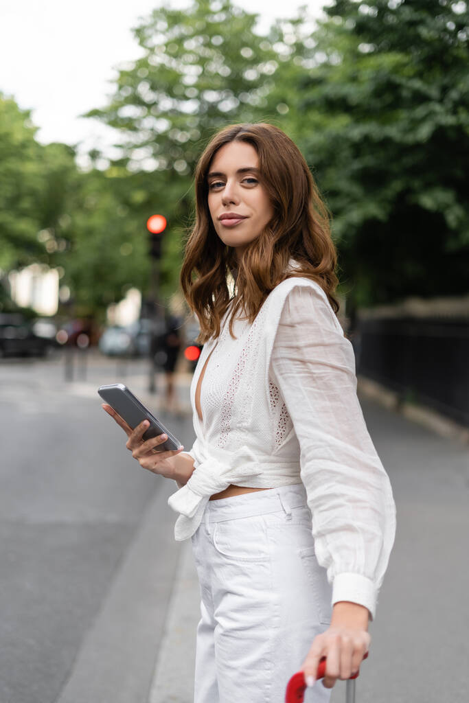Stílusos barna nő gazdaság smartphone és bőrönd az utcán Párizsban  - Fotó, kép