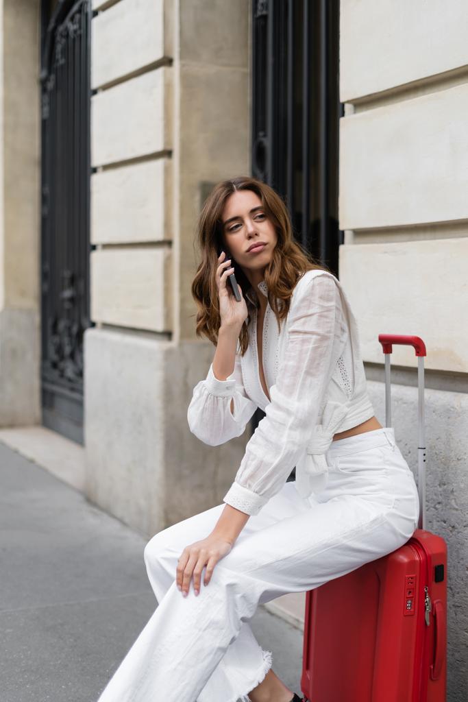Fashionable traveler talking on smartphone while sitting on baggage on street in Paris  - Photo, Image