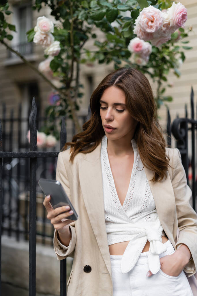Модная женщина в куртке с мобильным телефоном возле забора на улице в Париже  - Фото, изображение
