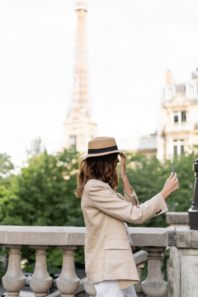 Vue latérale de la femme élégante en chapeau de soleil prenant selfie sur smartphone à Paris  - Photo, image