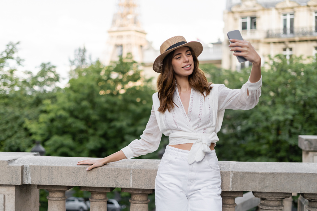 Wesoła kobieta w kapeluszu słonecznym robi selfie na smartfonie z zamazaną wieżą Eiffla w Paryżu  - Zdjęcie, obraz