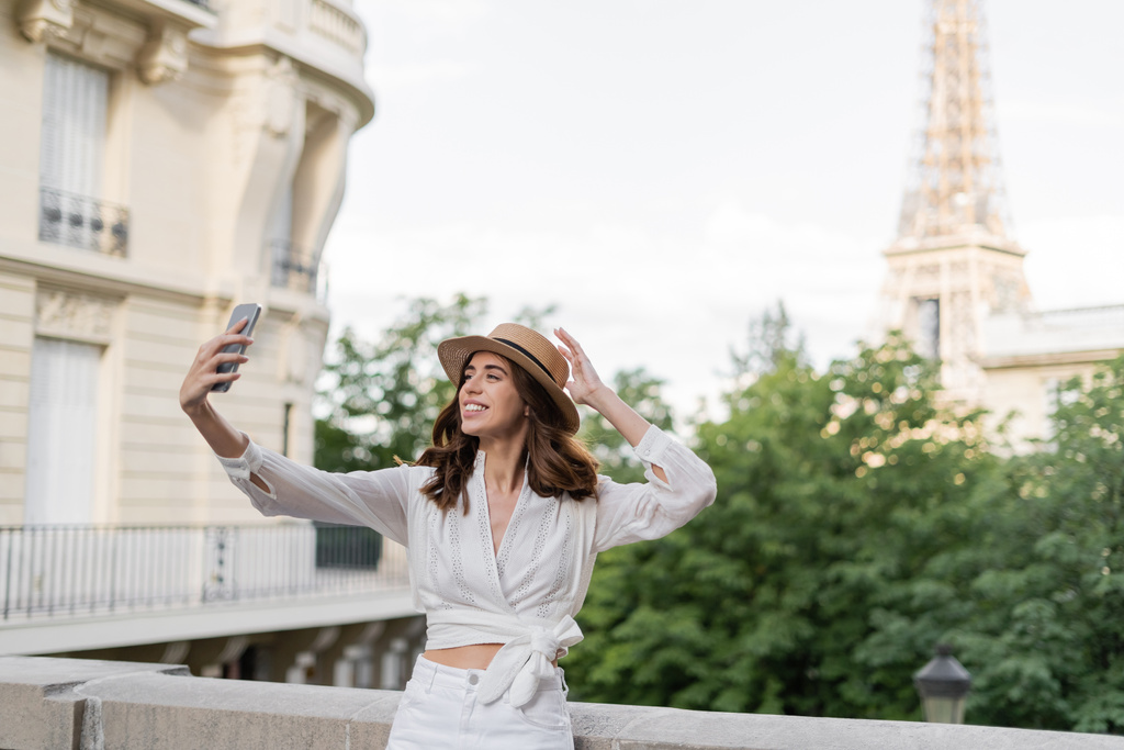 Positieve reiziger in zonnehoed selfie op smartphone met Eiffeltoren op de achtergrond in Frankrijk  - Foto, afbeelding