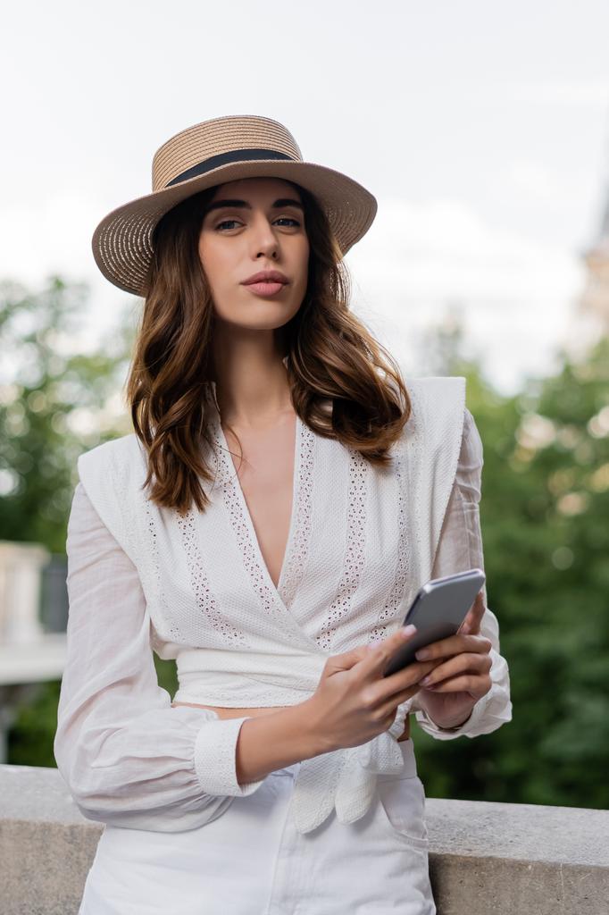 Retrato de la mujer de moda sosteniendo el teléfono inteligente y mirando a la cámara en la calle en París  - Foto, Imagen