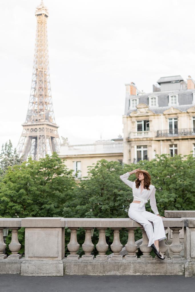 Elegante mujer sosteniendo sombrero de sol en la calle con la torre Eiffel al fondo en París  - Foto, imagen