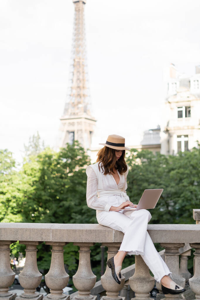 Güneş şapkalı şık bir kadın şehir sokağında dizüstü bilgisayar kullanıyor ve Paris 'te Eyfel kulesi var.  - Fotoğraf, Görsel