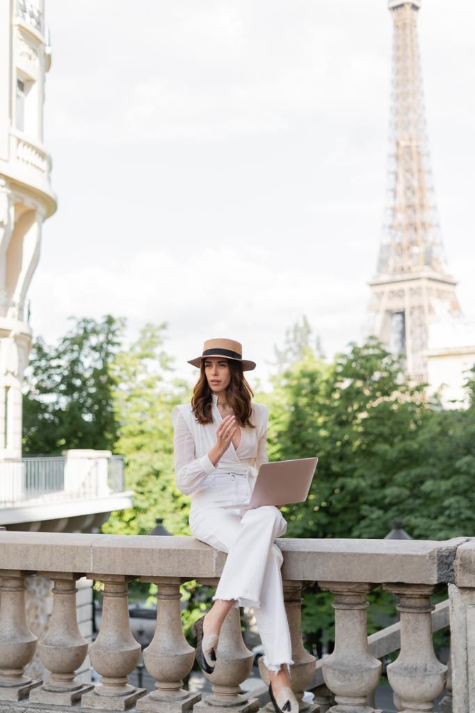 Trendige Frau mit Sonnenhut hält Laptop auf Straße mit Eiffelturm im Hintergrund in Paris  - Foto, Bild