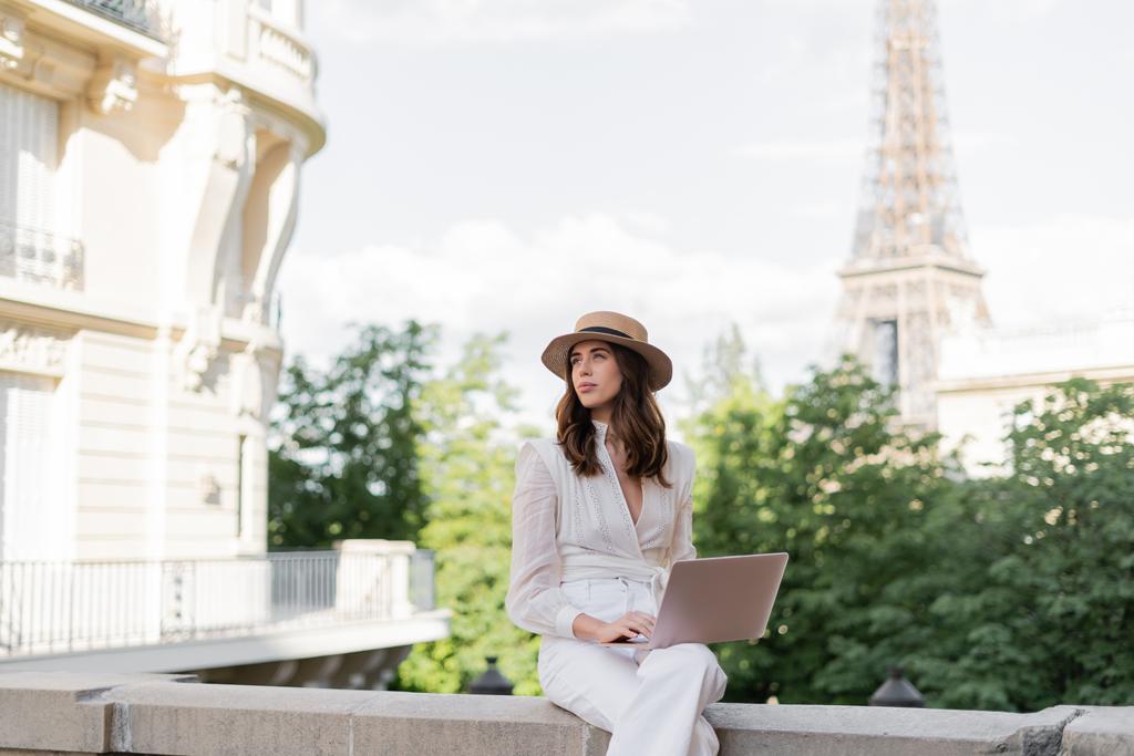 Modny stylowy freelancer w kapeluszu słonecznym przy użyciu laptopa na ulicy z wieżą Eiffla w tle w Paryżu  - Zdjęcie, obraz