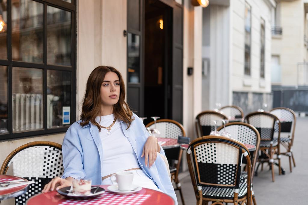 Femme élégante regardant loin tout en étant assis près du dessert et du café sur la table sur la terrasse du café à Paris  - Photo, image