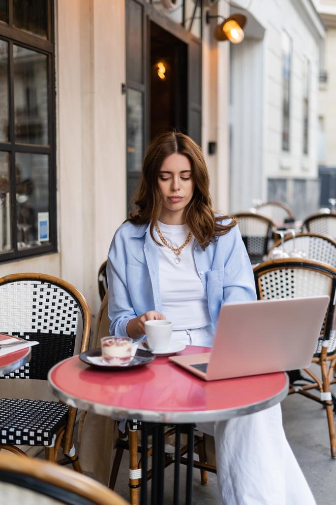 Freiberuflerin hält Kaffeetasse in der Nähe von Dessert und Laptop auf dem Tisch eines Cafés in Paris  - Foto, Bild