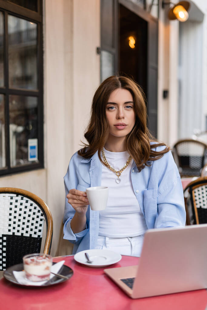 パリの屋外カフェでぼやけたデザートやノートパソコンの近くにコーヒーを持っているスタイリッシュな女性  - 写真・画像