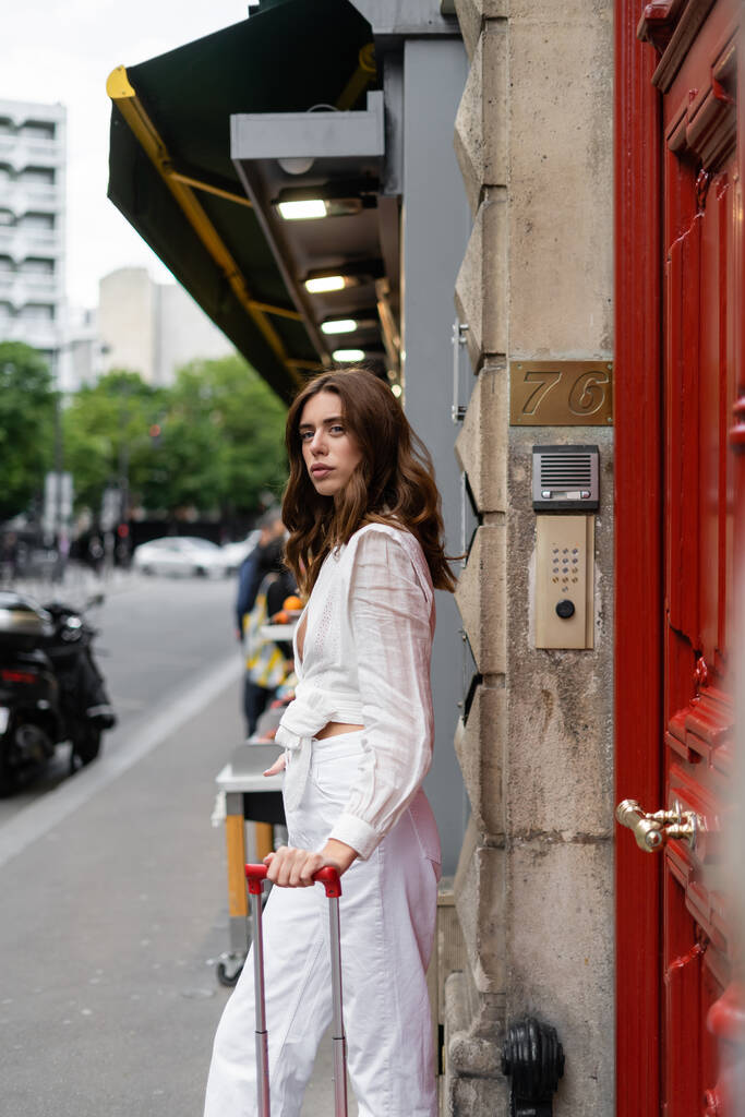 Stylowa kobieta stojąca przy walizce i budynku przy ulicy miejskiej we Francji  - Zdjęcie, obraz