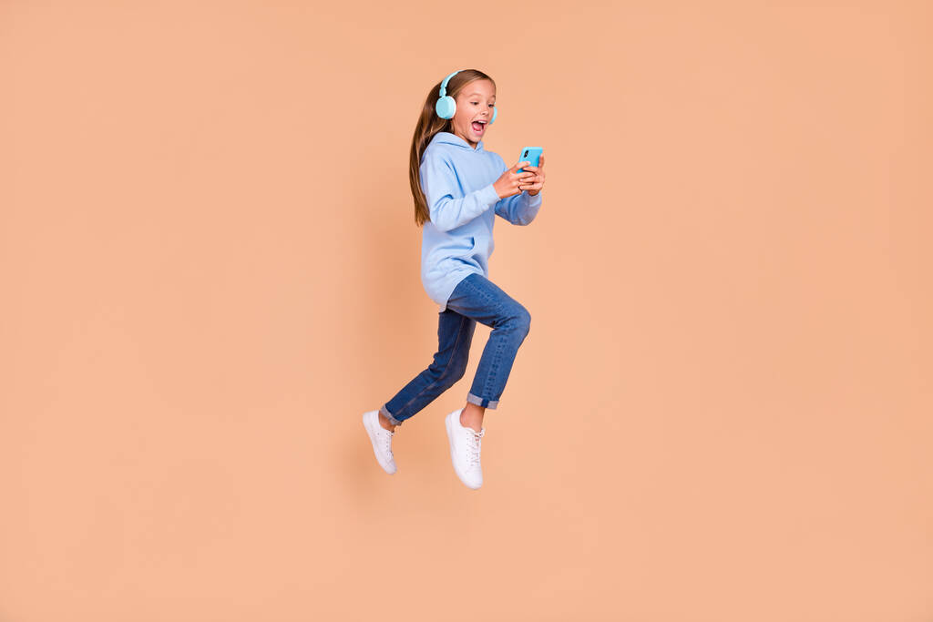 Heyecanlı güzel kız öğrencinin tam boy fotoğrafı atlama işlemi için bej renkli arka planda izole edilmiş telefon uygulaması kullan. - Fotoğraf, Görsel