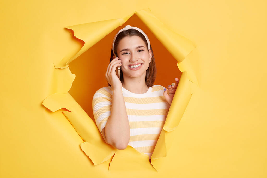 Εικόνα της χαμογελαστής χαρούμενης θετικής γυναίκας που φοράει ριγέ πουκάμισο και κορδέλα στα μαλλιά ποζάροντας σε κίτρινη χάρτινη τρύπα, μιλώντας μέσω έξυπνου τηλεφώνου, εκφράζοντας θετικά συναισθήματα, ακούγοντας καλά νέα. - Φωτογραφία, εικόνα