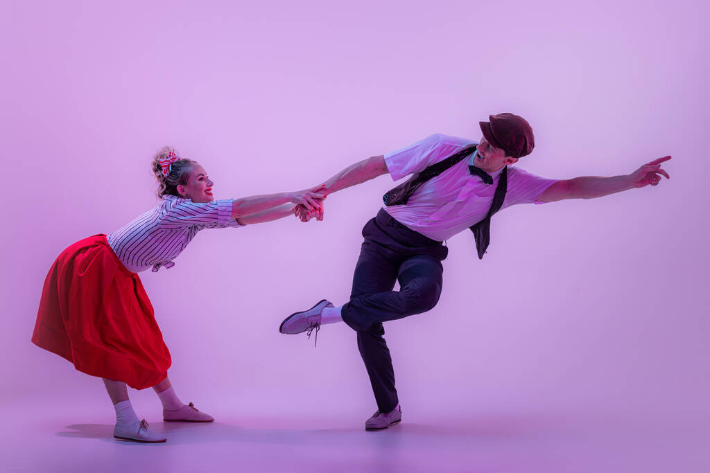 Společenský tanec. Studio záběr mladého muže a ženy ve vintage retro stylu oblečení tanec lindy hop izolované na šeříkové barvy pozadí v neonovém světle. Nadčasové tradice, módní styl a umění 60. let - Fotografie, Obrázek
