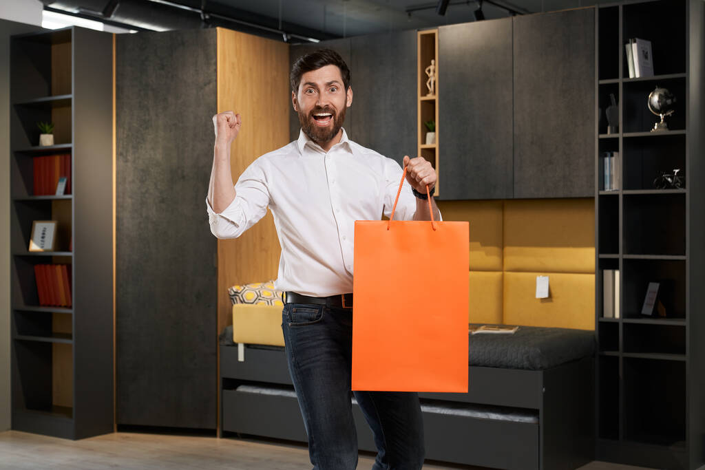 Excited, cheerful bearded man holding red shopping bag, while enjoying successful purchase in furniture store. Front view of happy guy raising hand up, while rejoicing shopping. Concept of emotions. - Photo, Image