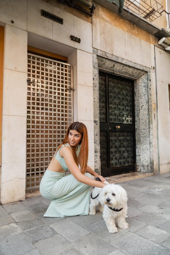 улыбающаяся женщина в стильном платье, смотрящая в сторону белой собаки на венецианской улице - Фото, изображение