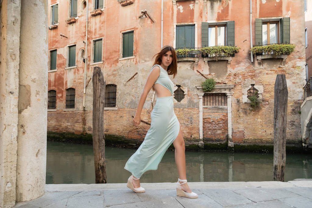 volledige lengte van trendy vrouw met rood haar wandelen langs kanaal op Venetiaanse straat - Foto, afbeelding