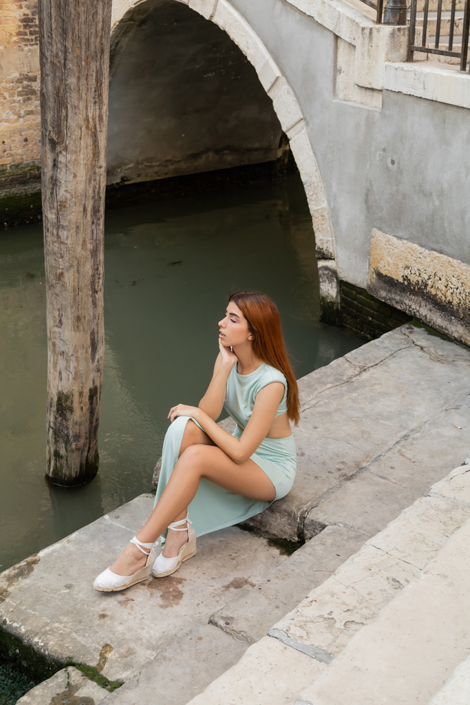 high angle view of stylish woman with red hair sitting on stairs near urban venetian canal - 写真・画像