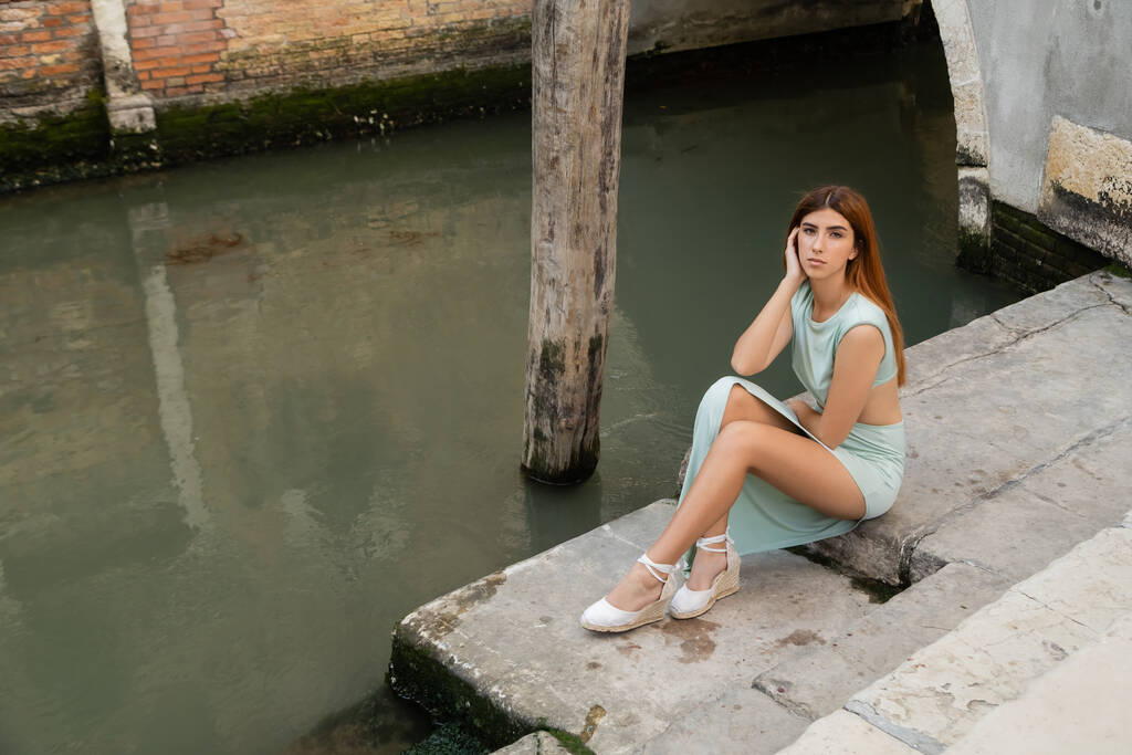 όμορφη γυναίκα κοιτάζει κάμερα, ενώ κάθεται σε σκάλες κοντά στο κανάλι στη Βενετία - Φωτογραφία, εικόνα