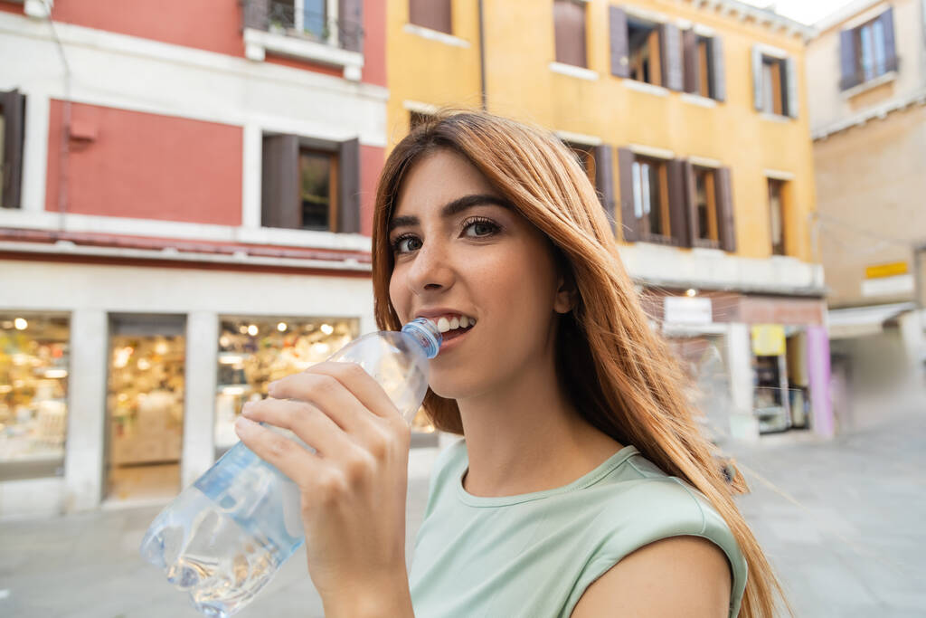 happy young woman with bottle of water looking at camera on blurred venetian street - 写真・画像