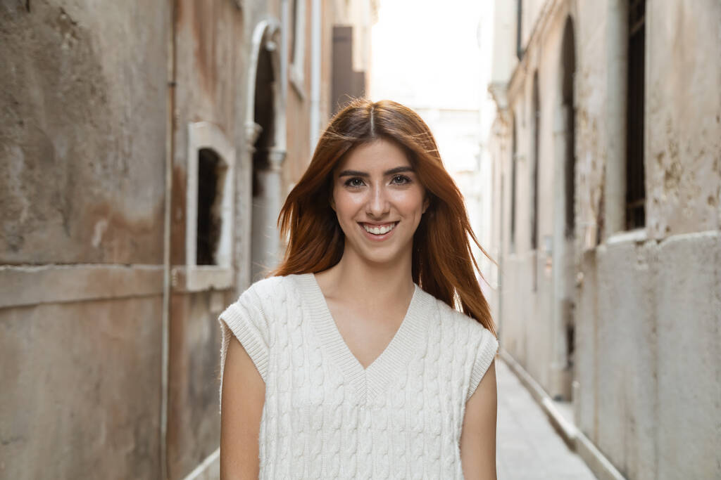 retrato de mujer encantadora y feliz con el pelo rojo cerca de edificios borrosos en Venecia - Foto, imagen