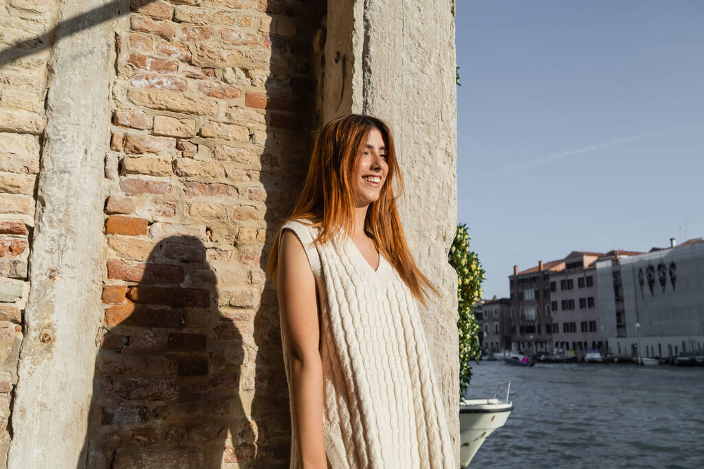 cheerful redhead woman in sleeveless jumper smiling near stone wall in Venice - Photo, Image