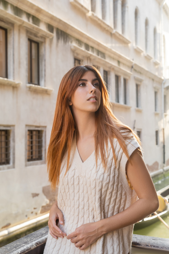 jonge roodharige vrouw kijkt weg in de buurt van wazig middeleeuws gebouw in Venetië - Foto, afbeelding